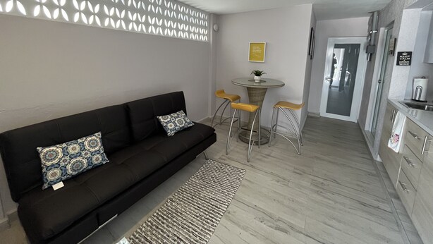 Un salon moderne avec un canapé noir, des coussins à motifs, une table ronde avec des tabourets jaunes, un panneau décoratif mural et un coin cuisine équipé d’un évier et de placards.