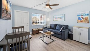 A modern living room with a sofa, a dining table, a ceiling fan, and artwork on the walls. - Madeira Beach Getaway – Tropic Breeze 7 Pool View Condo (Madeira Beach)