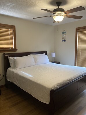 A neatly made bed in a room with wooden flooring, a ceiling fan, and a wall-mounted light fixture.