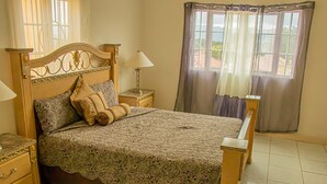 A bedroom with a wooden bed, lace bedspread, two bedside tables with lamps, and a view of the outdoors through a window with curtains. - HOLIDAYS AT MANGO WALK COUNTRY CLUB (Montego Bay)