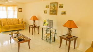 A living room with a yellow sofa, two lamps, a coffee table, and a TV stand. - HOLIDAYS AT MANGO WALK COUNTRY CLUB (Montego Bay)