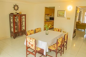 A dining area with a table set for six, surrounded by floral-patterned chairs. A wooden cabinet with glass doors is to the left, and a kitchen is visible in the background.