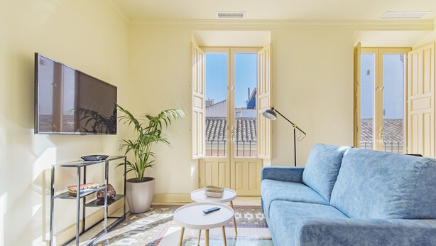 Una sala de estar con un sofá, un televisor, una mesita, una planta y una ventana con puerta abierta.