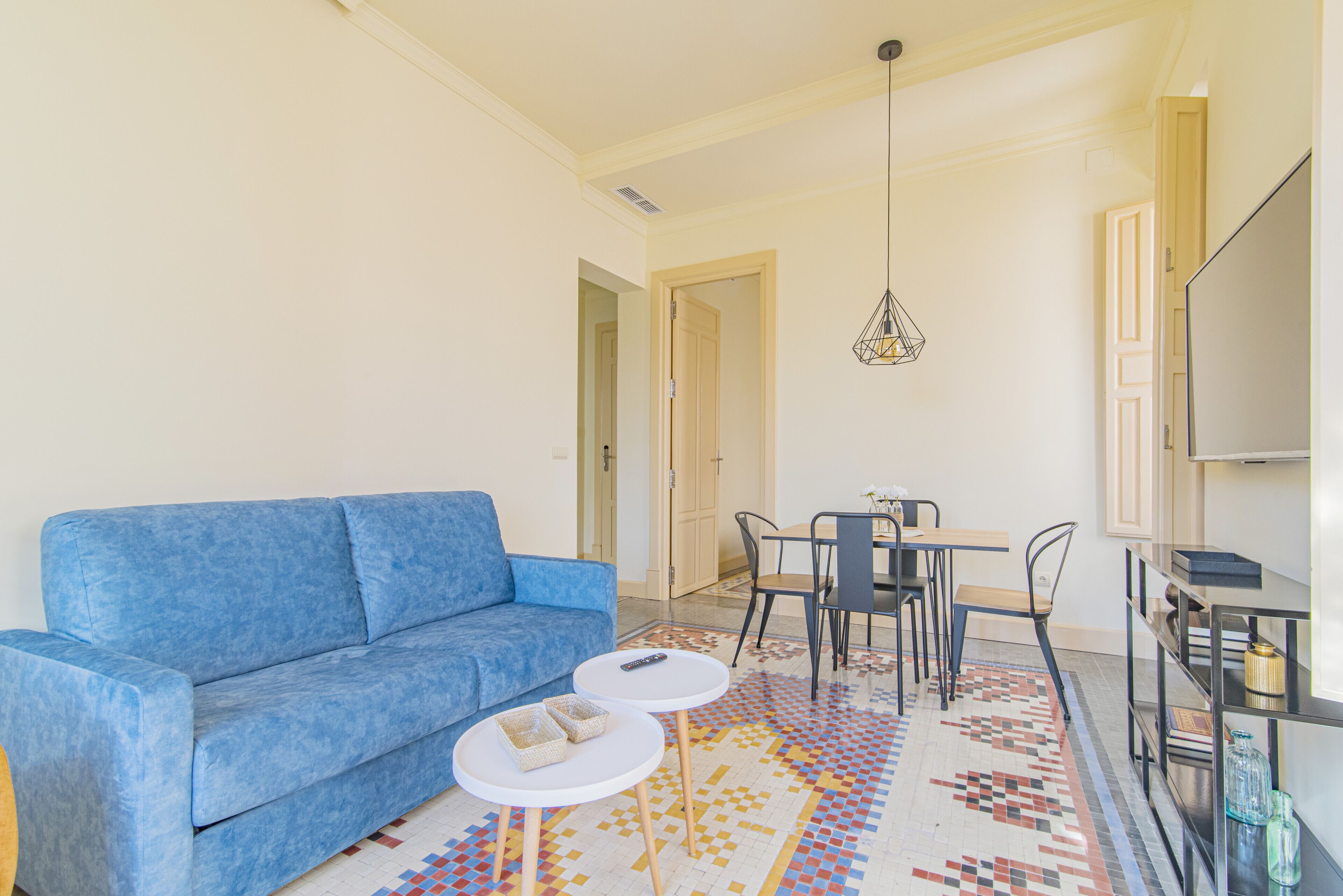 Una sala de estar con un sofá azul, una mesita redonda con un control remoto, una alfombra estampada y un televisor sobre un mueble.