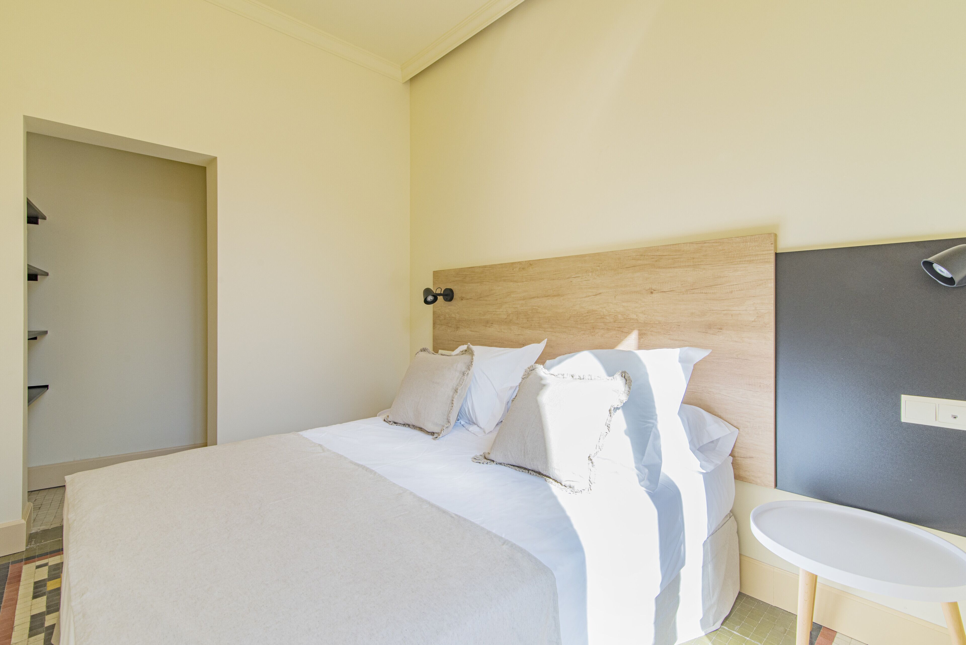 A neatly made bed with white linens and pillows, a wooden headboard, a bedside table, and a small white sink.