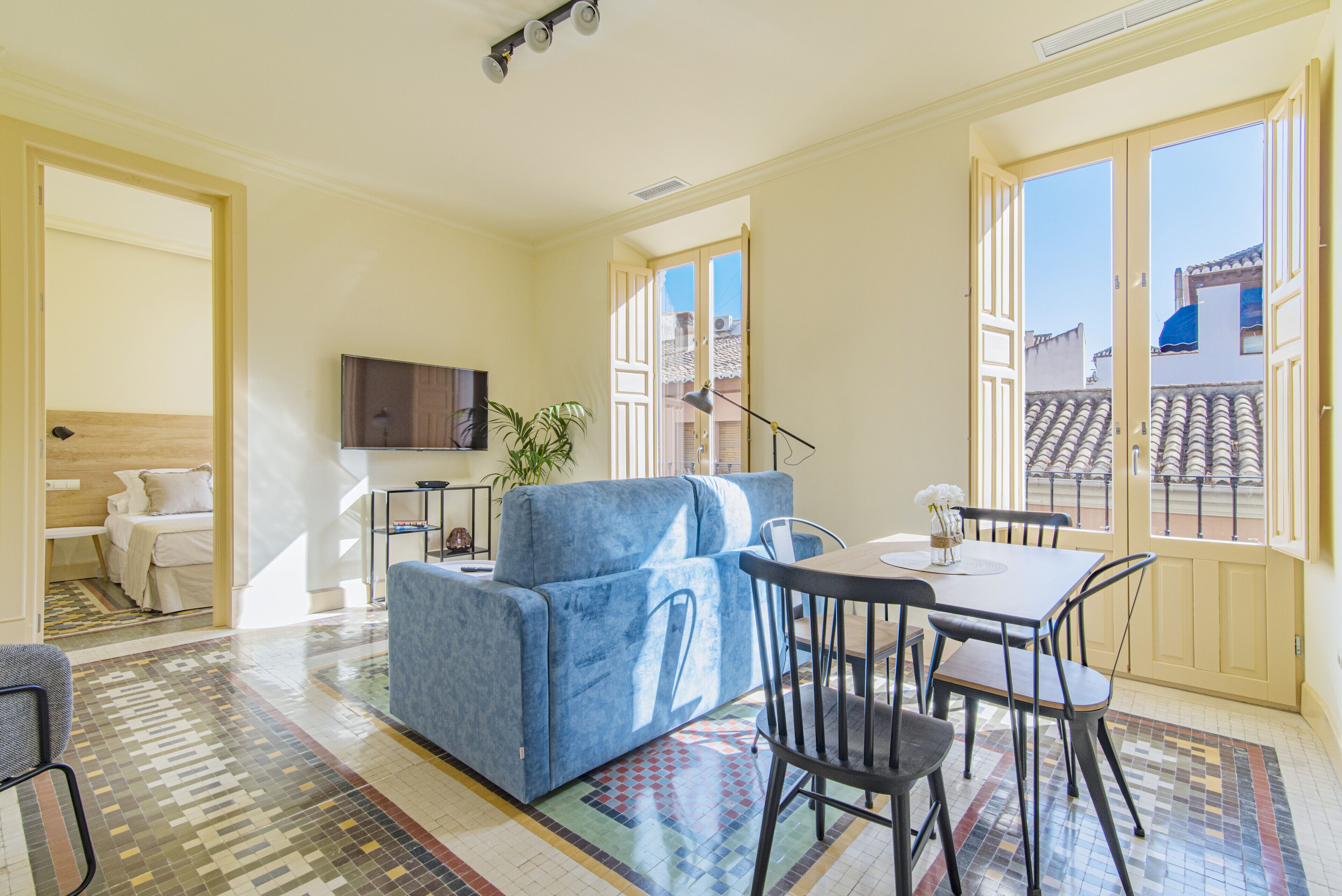 A modern living room with a blue sofa, dining table, and chairs, a flat-screen TV, and large windows with a view of buildings.