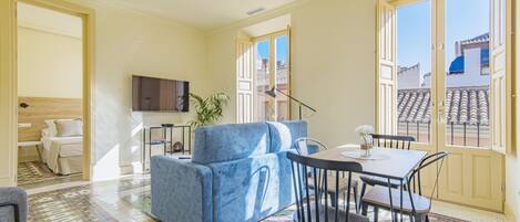 A modern living room with a blue sofa, dining table, and chairs, a flat-screen TV, and large windows with a view of buildings.