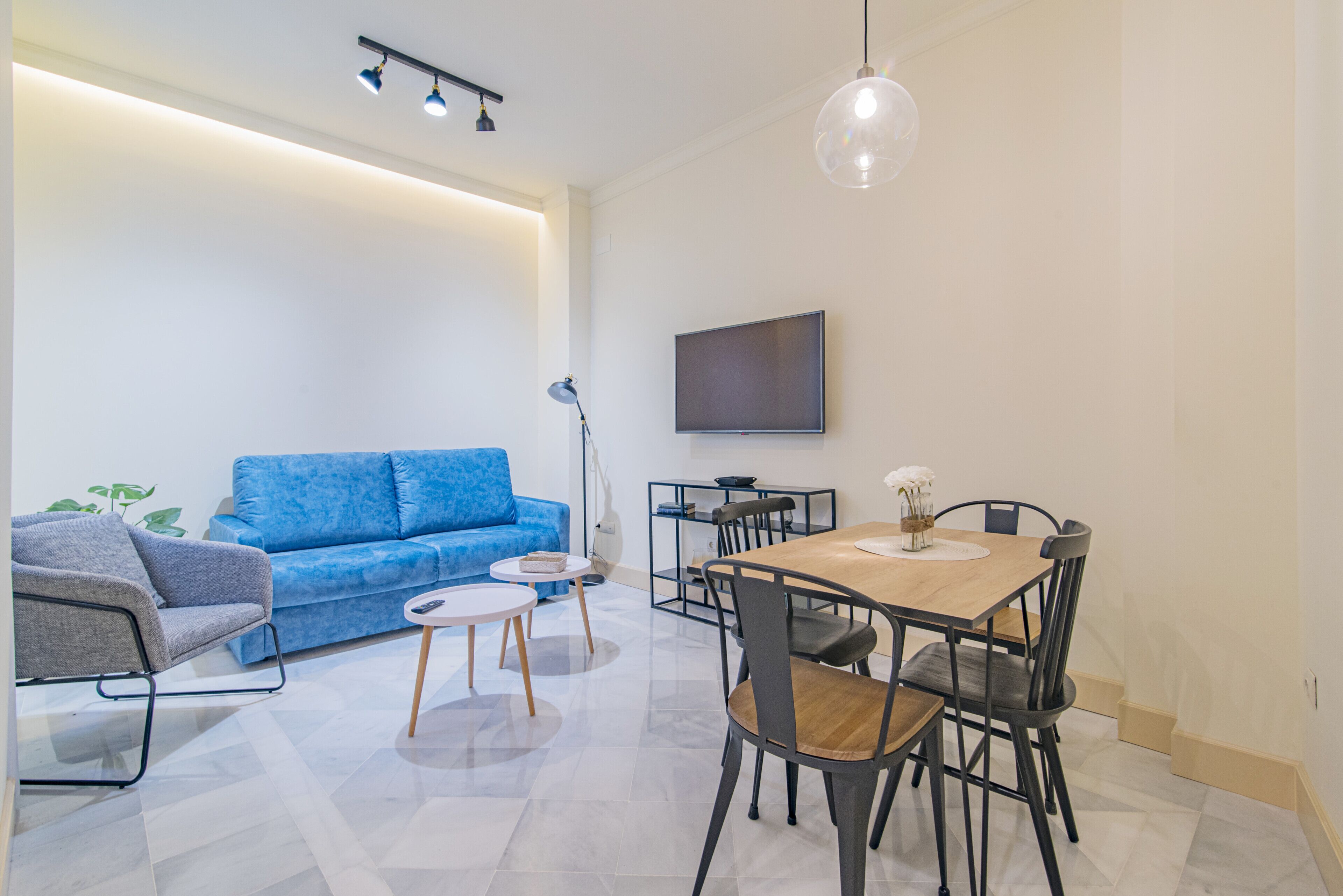 Een moderne woonkamer met een blauwe bank, een eettafel met stoelen, een aan de muur bevestigde televisie en een hanglamp.