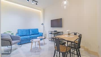 Een moderne woonkamer met een blauwe bank, een eettafel met stoelen, een aan de muur bevestigde televisie en een hanglamp.