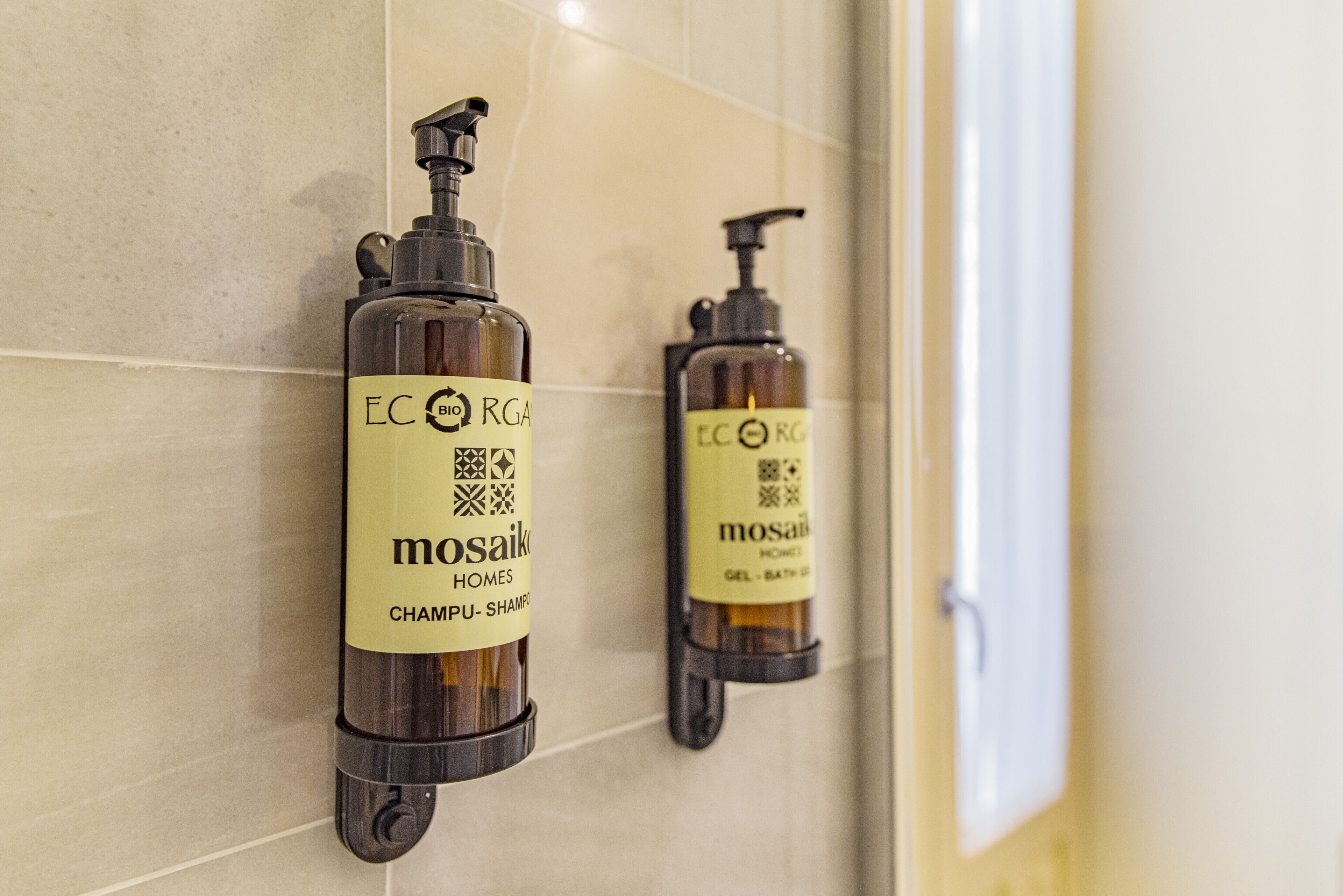 Two wall-mounted shampoo dispensers labeled 'mosaiK HOMES CHAMPU-SHAMPO' on a tiled wall.