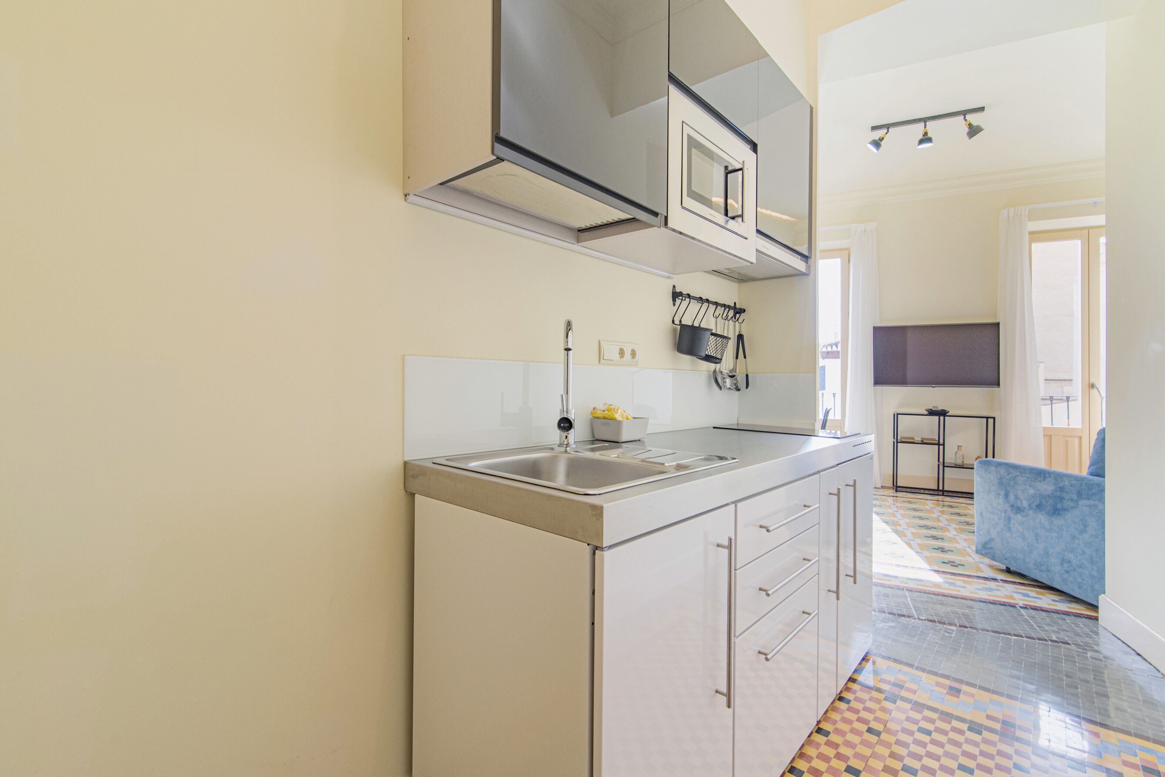 A modern kitchen with a stainless steel sink, white cabinets, and a microwave. There is a patterned floor and a blue chair in the adjacent room.