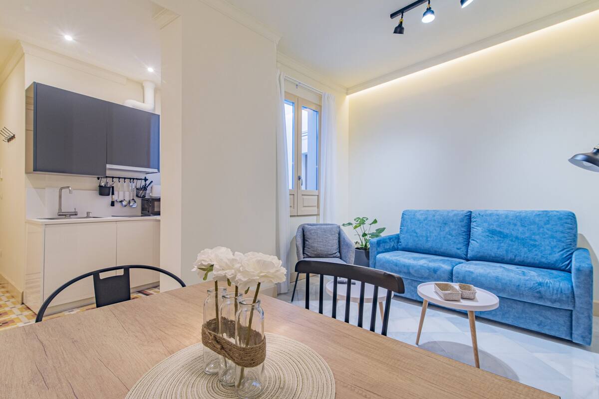 Una sala de estar moderna con un sofá azul, una mesa de comedor de madera con un jarrón de flores blancas, y una zona de cocina al fondo.