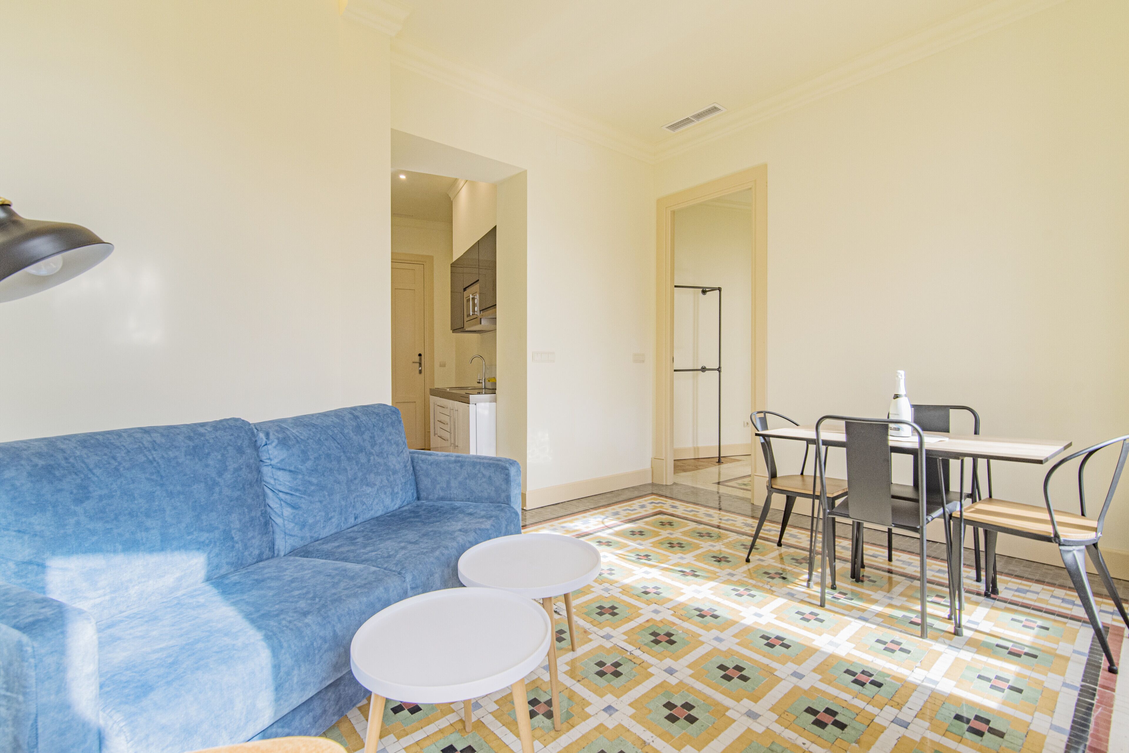 Una sala de estar con un sofá azul, dos mesitas blancas, una mesa de comedor con sillas y un piso con estampado.