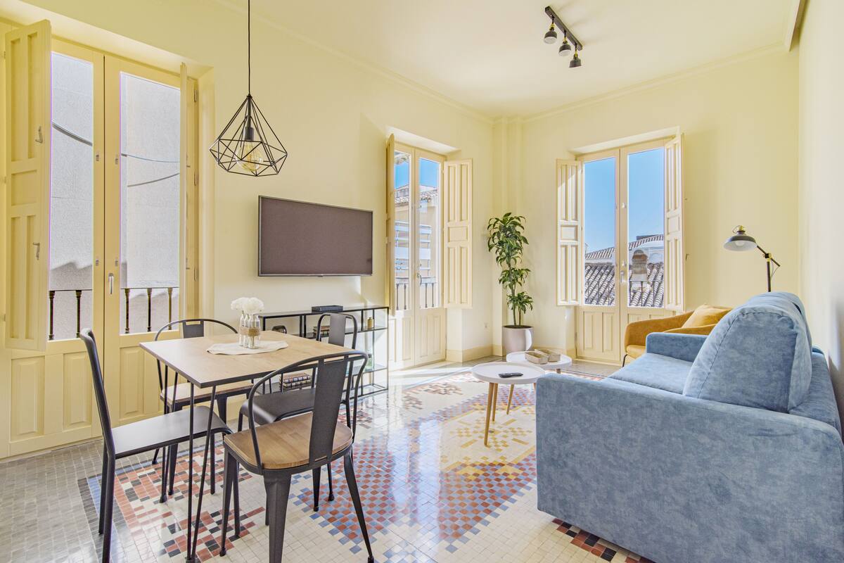 Amplia sala de estar con sofá, mesa de comedor y sillas, televisor montado en la pared y una planta en maceta.