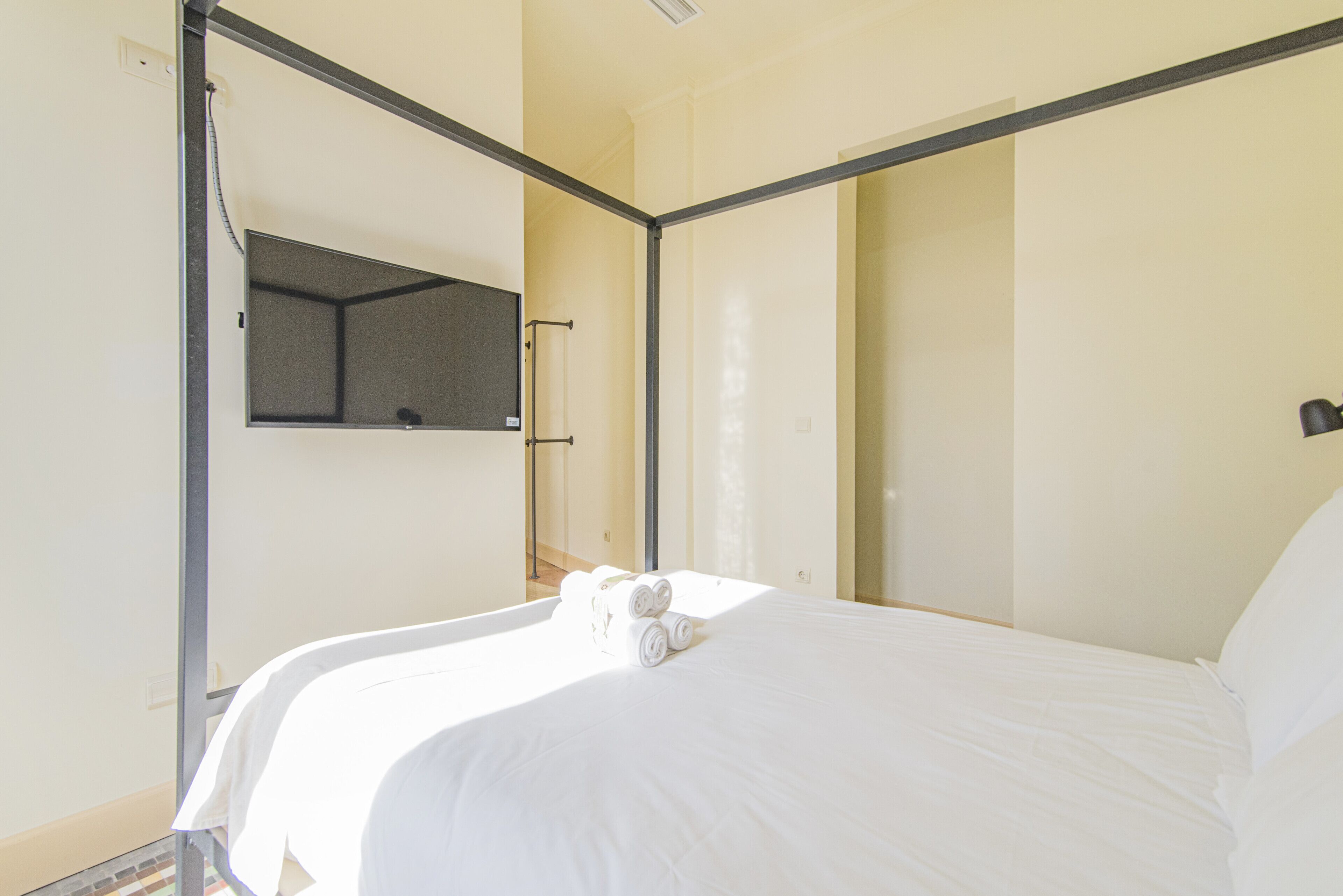 A single bed with white linens and rolled towels, a bedside table, and a wall-mounted TV.