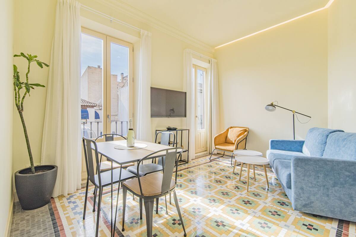 Una sala de estar con un sofá azul, una mesa de comedor con sillas, una planta en maceta y un televisor sobre un mueble.