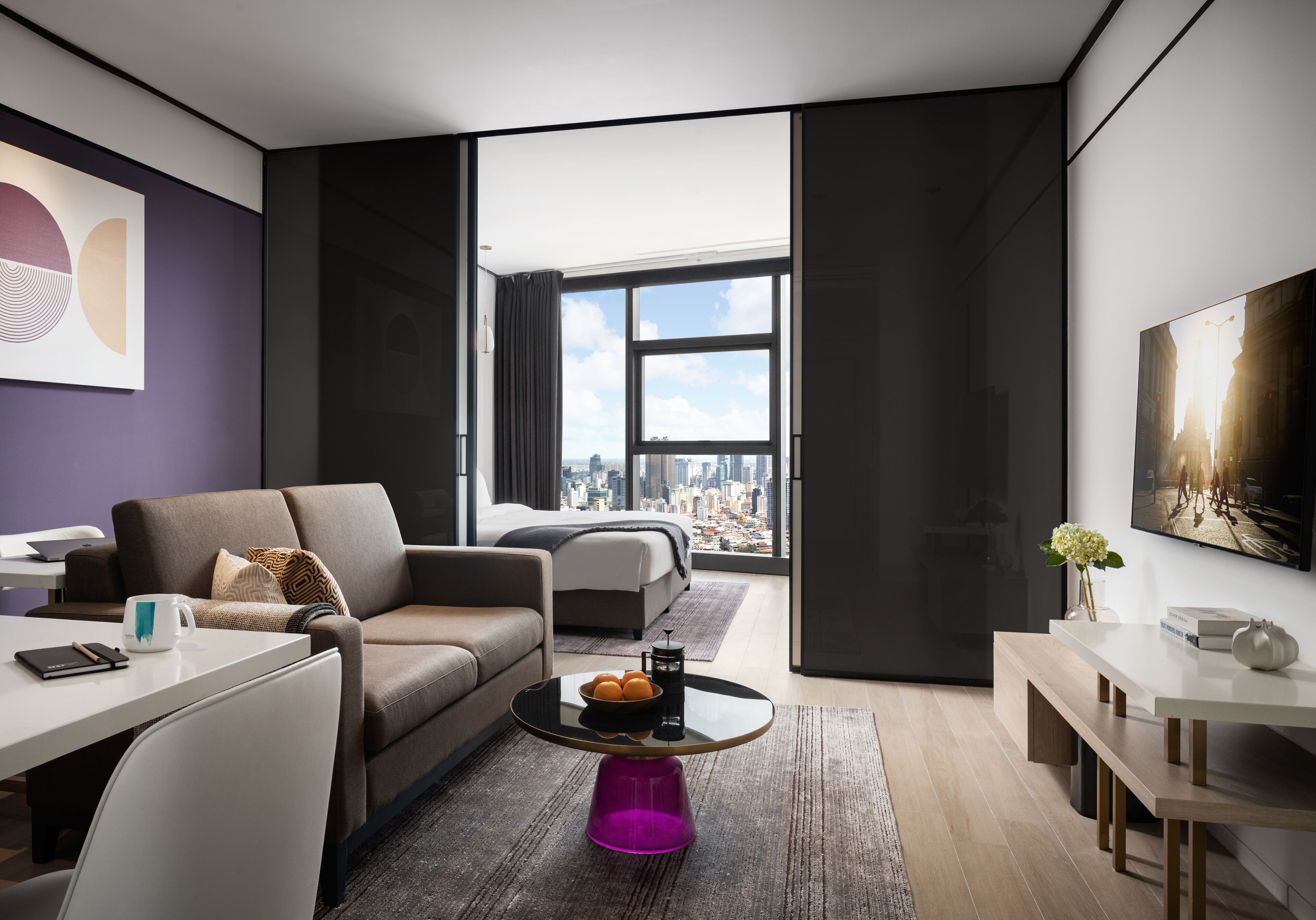 A modern hotel room with a city view, a sofa, a coffee table with oranges, and a dining area.