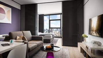 A modern hotel room with a city view, a sofa, a coffee table with oranges, and a dining area.