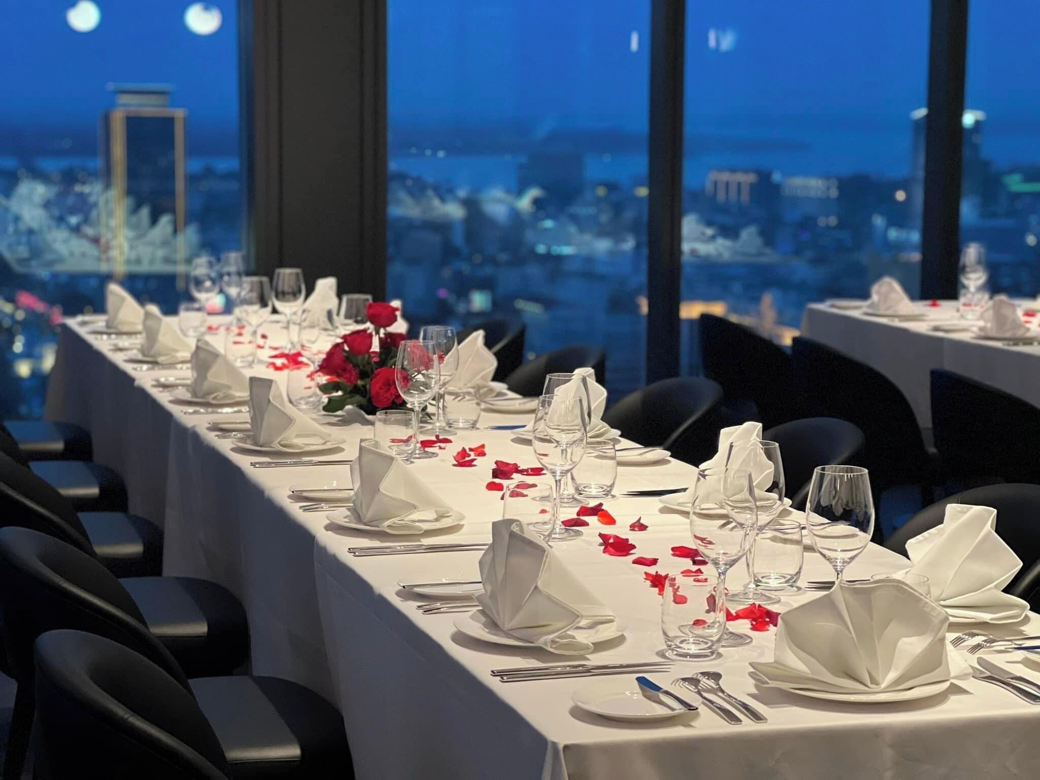 A long dining table set for a formal event, with white tablecloths, folded napkins, and rose petals.