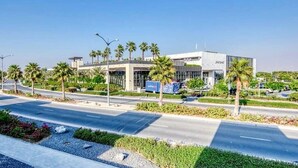 A modern building with a flat roof, surrounded by palm trees and a landscaped area with a road in front. - Luxurious 3 Bedrooms with Maid room Villa (دبي)