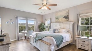 Une chambre à coucher avec un lit, des tables de chevet, un bureau et une chaise. On y trouve un ventilateur de plafond, une fenêtre avec des rideaux et un tableau accroché au mur.