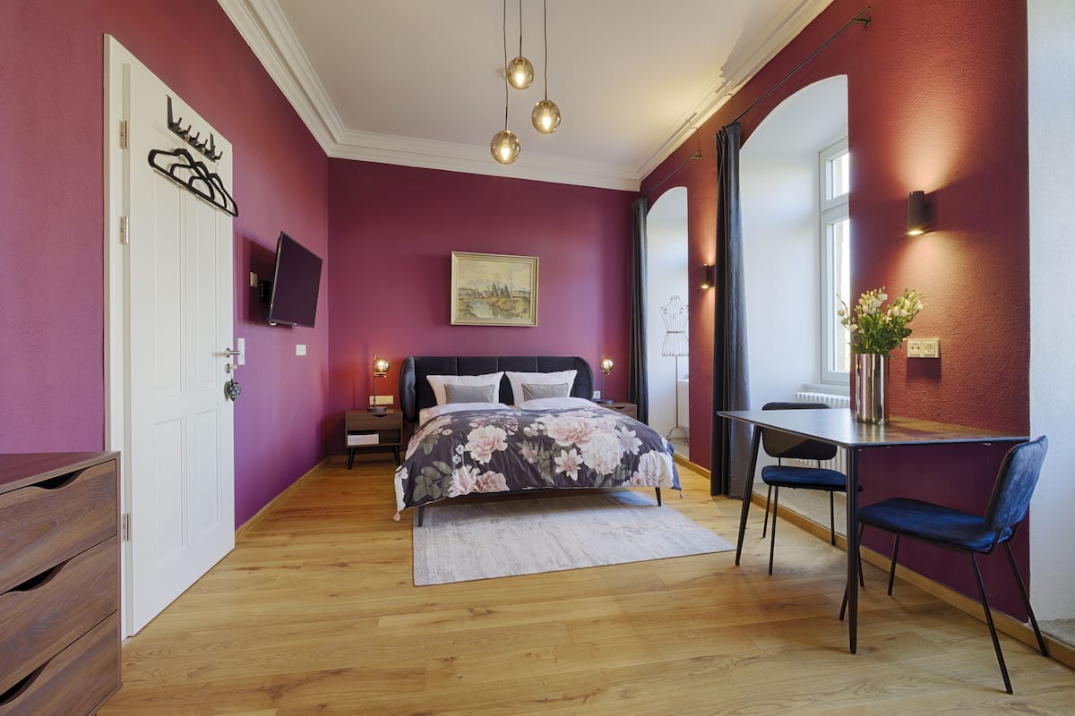 Une chambre à coucher avec un lit, un bureau et une chaise. Il y a un téléviseur accroché au mur et une fenêtre avec des rideaux.
