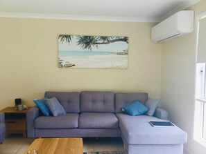 A living room with a grey sofa, two blue cushions, a wooden coffee table, and a beach-themed painting on the wall. - Tides Inn with Plunge Pool (Agnes Water)