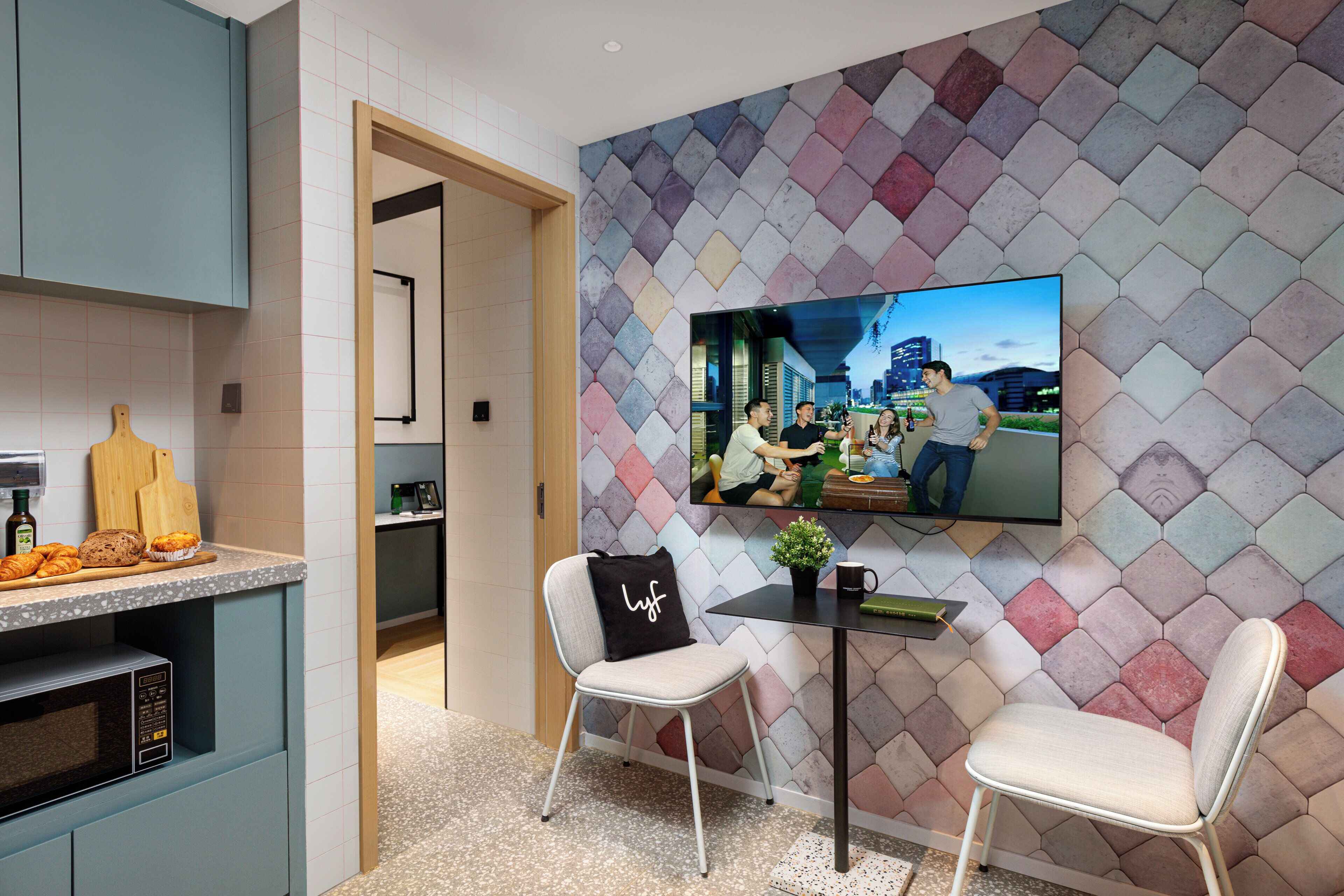 A modern kitchen with a microwave, a cutting board, and a loaf of bread. There is a TV mounted on the wall displaying a cityscape. Two white chairs are placed in front of a small table with a plant and a book.