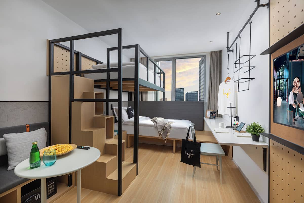 A compact hotel room with a bunk bed, a small kitchenette, a dining area, and a large window offering a city view.
