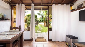 Une pièce avec une table en bois, des chaises, un banc, une poubelle et une vue sur un jardin avec un sentier et des plantes.