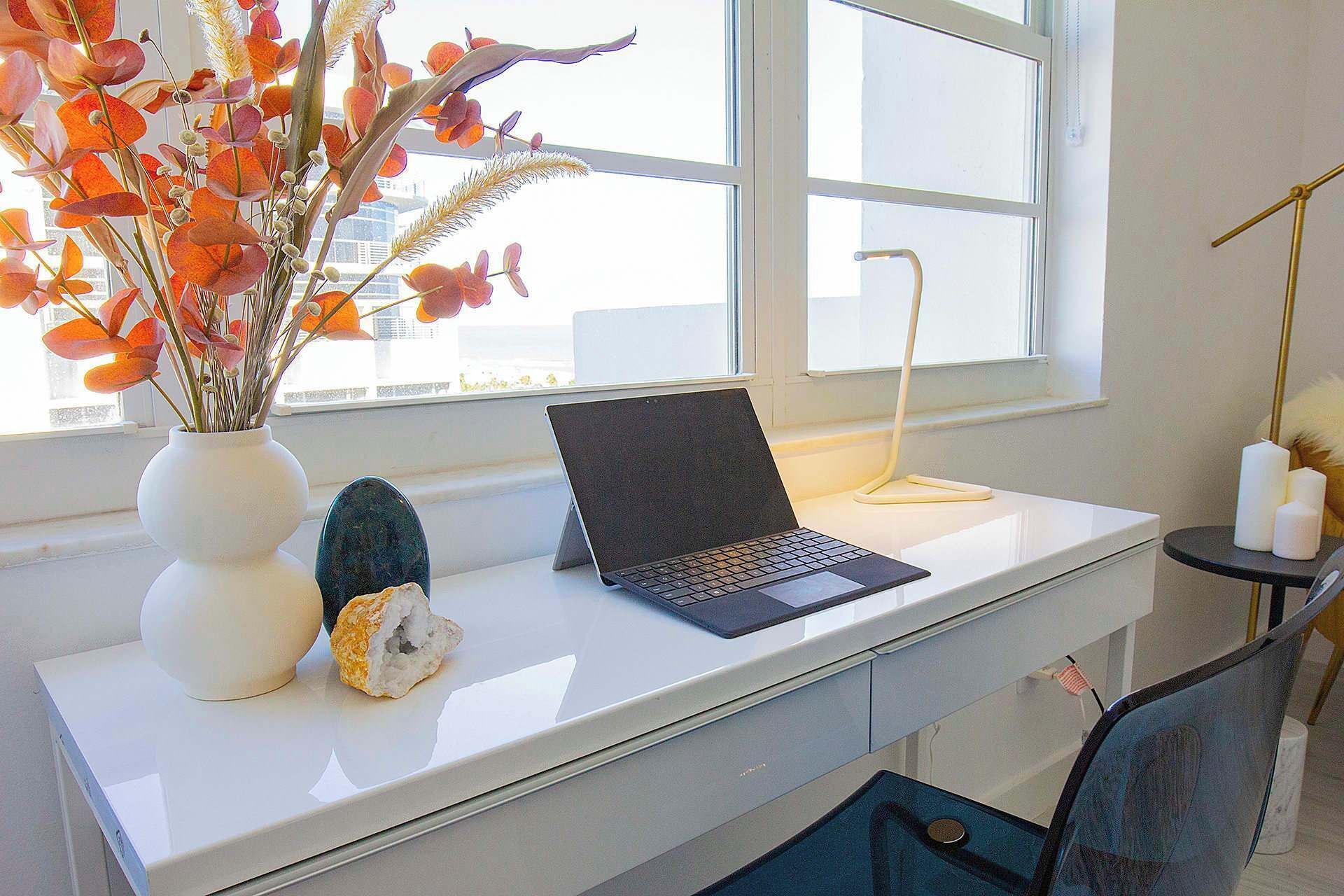 Un espacio de trabajo con una computadora portátil sobre un escritorio blanco, un florero con flores y una pequeña planta.