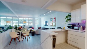 A modern kitchen with a marble island, dining area with chairs, and a living space with a sofa and a TV. - NEW! Luxury Ocean View  – 1Bd 2Bth – Designer Made & Breathtaking Views (Miami Beach)