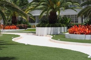 A landscaped area with palm trees and ornamental plants, a curved pathway, and a building with large windows in the background.