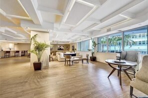 A spacious, well-lit room with a wooden floor, a round table, and a sofa. There is a large window providing a view of palm trees and a building outside.