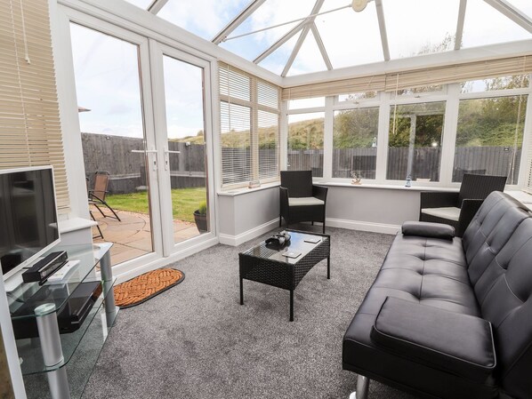 A conservatory with a glass ceiling, a black leather sofa, a glass coffee table, and a TV on a stand.