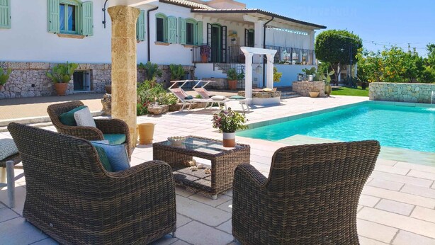 A poolside area with wicker furniture, a stone wall, and a building with green shutters.