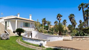 A white villa with a curved driveway, palm trees, and a stone wall.
