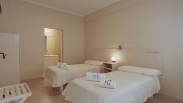 A hotel room with two beds, each with a white bedspread and a towel with 'HT' on it. There is a small table and a nightstand with a lamp.