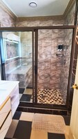 Un baño con cabina de ducha de vidrio, paredes de mármol, piso a cuadros y lavamanos blanco.