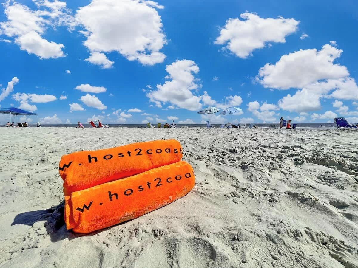 Una spiaggia sabbiosa con due asciugamani arancioni, con le scritte 'host2coast' e 'w.host2coast'.