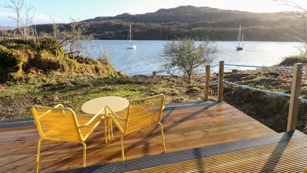 A wooden deck with yellow chairs and a table overlooking a lake with sailboats. - Kelpie (Lochgilphead)