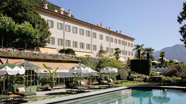 Un área de piscina con sillones de descanso y sombrillas, un edificio grande con múltiples ventanas y un paisaje montañoso de fondo.