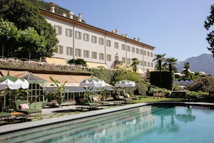 Un área de piscina con sillones de descanso y sombrillas, un edificio grande con múltiples ventanas y un paisaje montañoso de fondo.