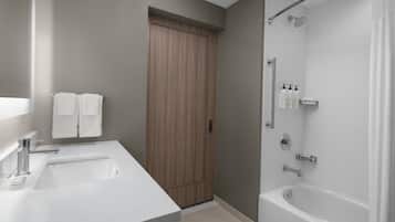 Un baño con lavamanos, espejo, puerta de madera, ducha y artículos de aseo en un estante.
