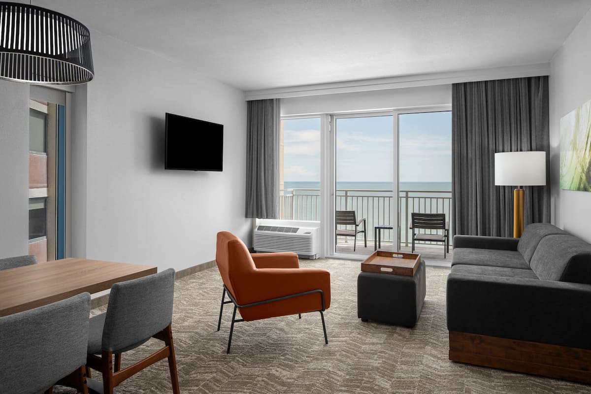 A hotel room with a wooden dining table, a sofa, a chair, a TV, and a balcony with a view of the sea.