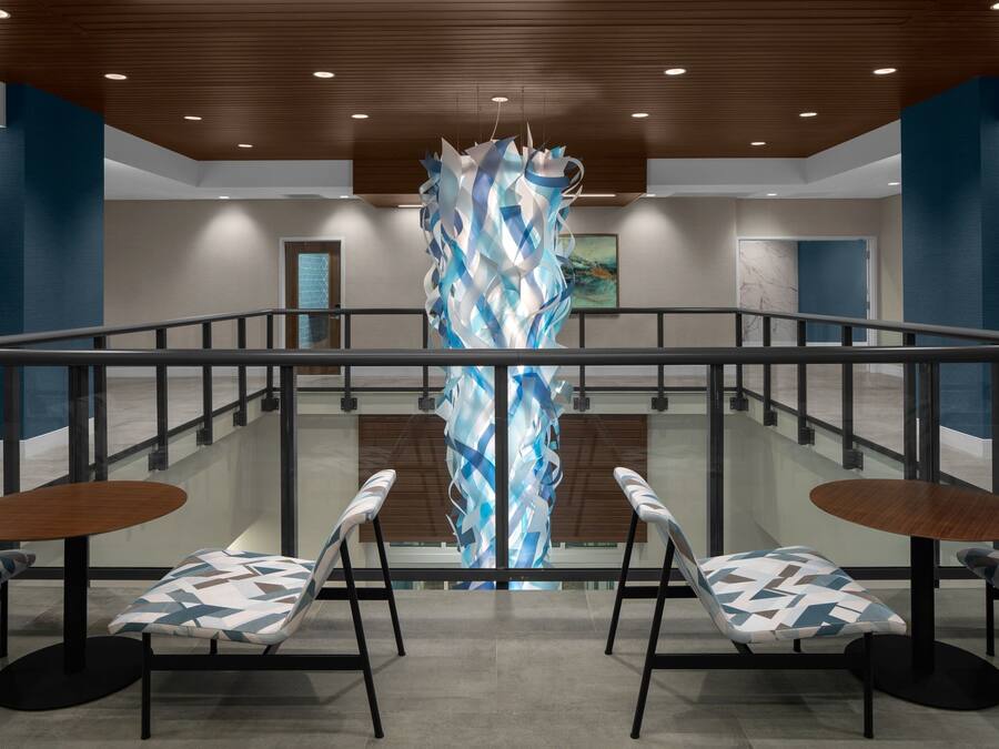 A modern lobby with a central abstract sculpture, two round tables with patterned chairs, and a glass railing.