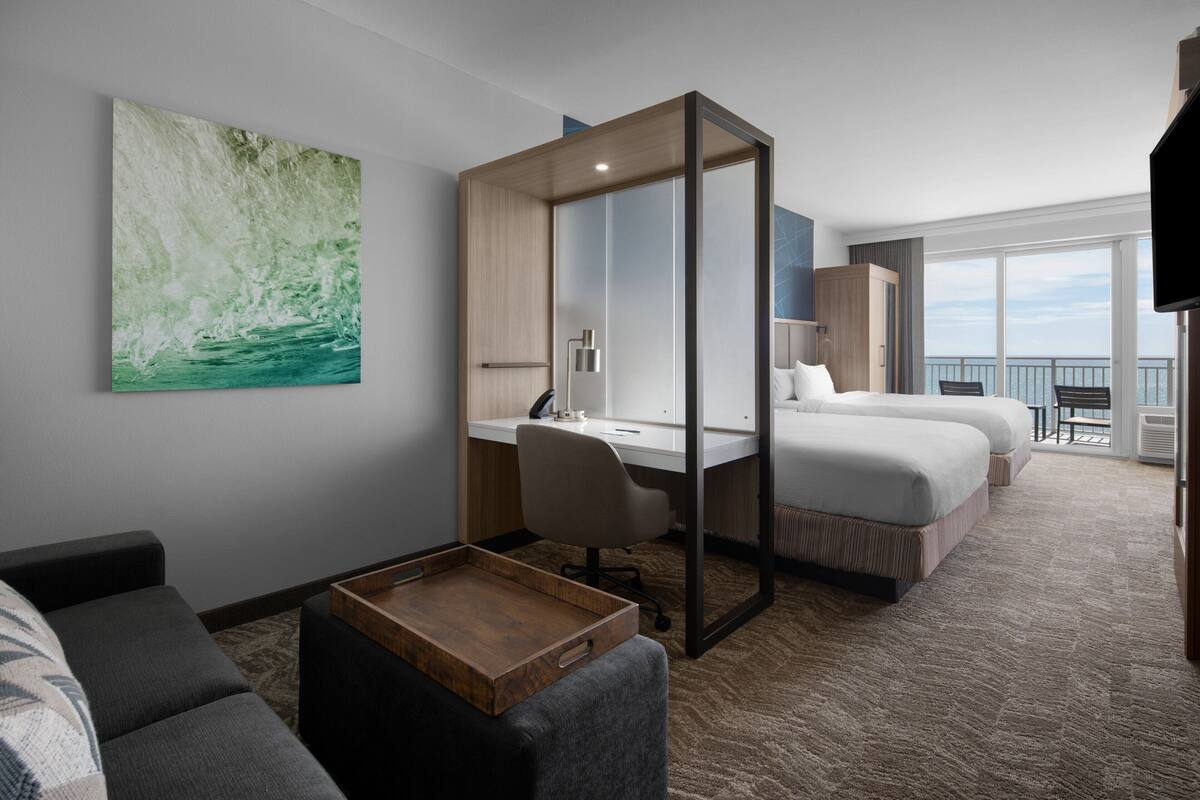 A hotel room with a desk, two beds, a large window with a view, a TV, and a painting on the wall.