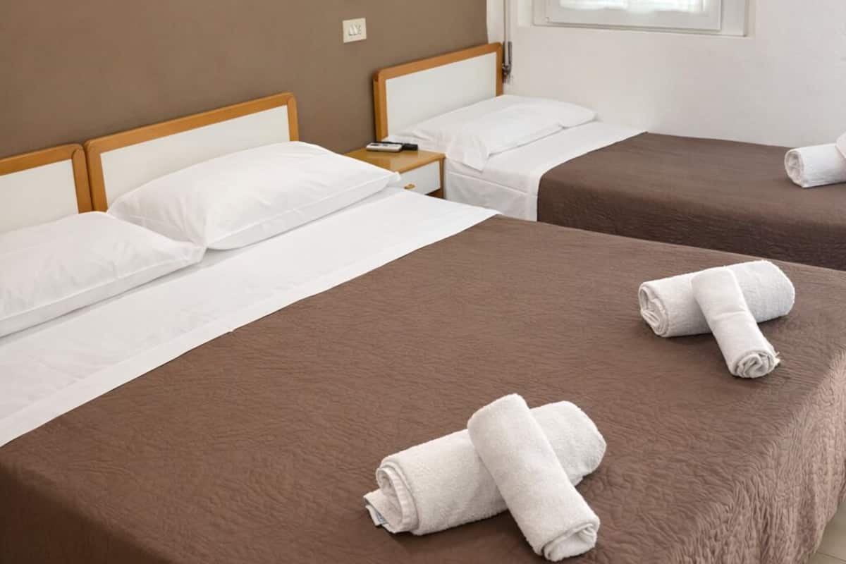 A hotel room with two beds, each with a brown bedspread and white linens. There is a framed picture on the wall, a bedside lamp, and a window with blinds.