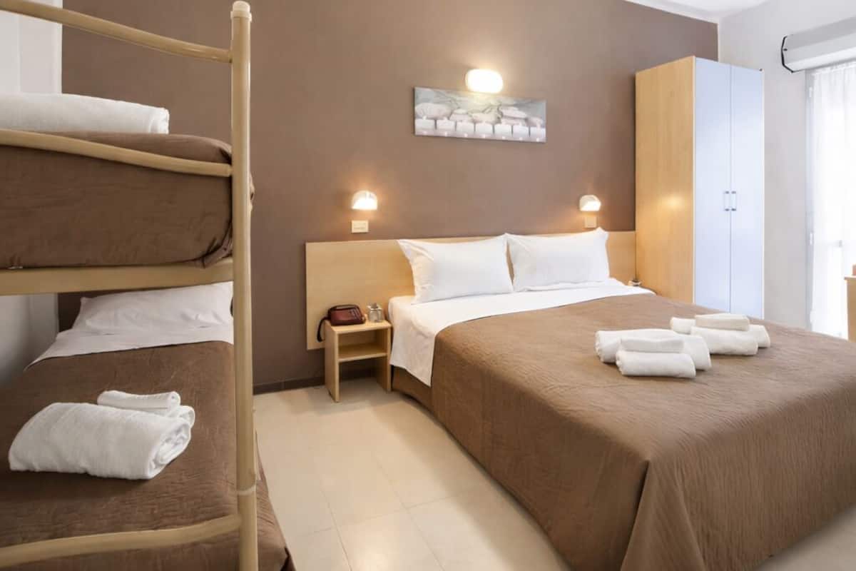 A hotel room with a bunk bed, a single bed, a nightstand, a lamp, a wardrobe, and a painting on the wall.