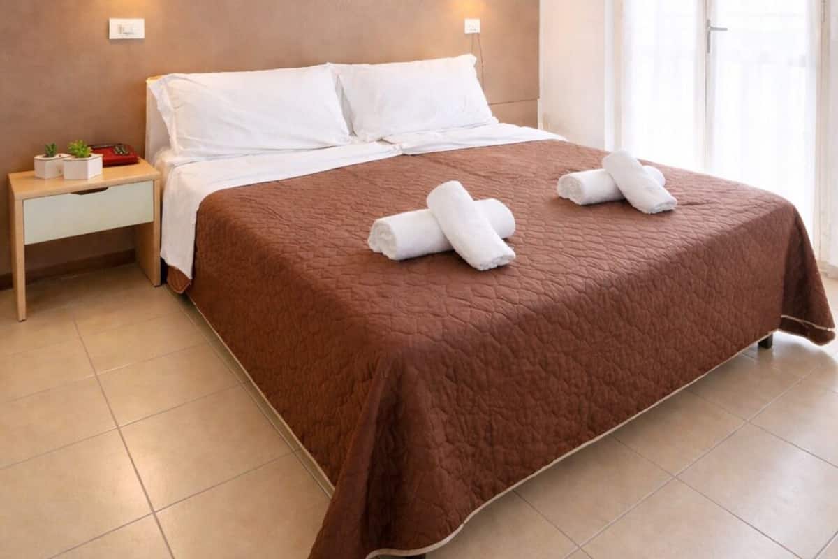 A neatly made bed with a brown bedspread and white pillows, two wall-mounted lights, and a window with sheer curtains.